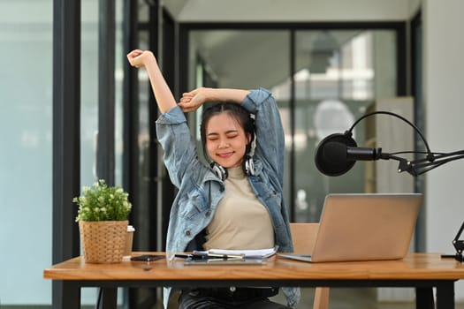 Carefree Asian female radio host relaxing on chair after recording podcast at home studio..