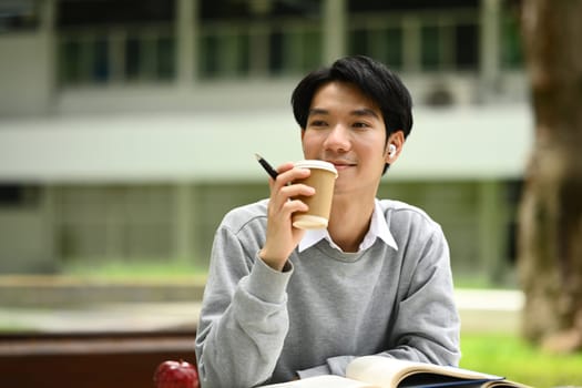 Carefree asian university student man drinking coffee and relaxing in the park on beautiful day.