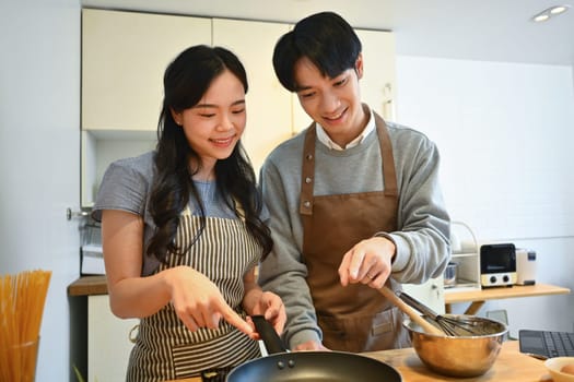 Happy young Asian couple on aprons making pancakes for breakfast, spending a weekend morning together.