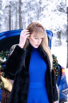 Woman in winter snowy forest in blue dress next to blue car decorated with Christmas decor. Christmas and winter holidays concept