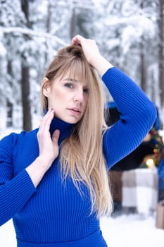 Woman in winter snowy forest in blue dress next to blue car decorated with Christmas decor. Christmas and winter holidays concept