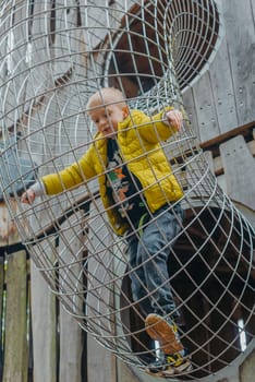 A child climbs up an alpine grid in a park on a playground on a hot summer day. children's playground in a public park, entertainment and recreation for children, mountaineering training. Child playing on outdoor playground. Kids play on school or kindergarten yard. Active kid on colorful slide and swing. Healthy summer activity for children. Little boy climbing outdoors.