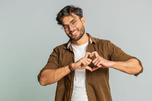 Man in love. Smiling attractive Indian man makes heart gesture demonstrates love sign expresses good positive feelings and sympathy. Handsome Arabian Hindu young guy isolated on gray wall background