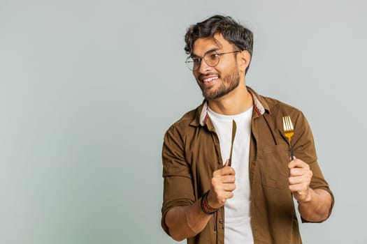 Ready to eat. Hungry Indian man waiting for serving dinner dishes with with restlessness holding cutlery fork knife will appreciate delicious restaurant meal. Excited guy isolated on gray background