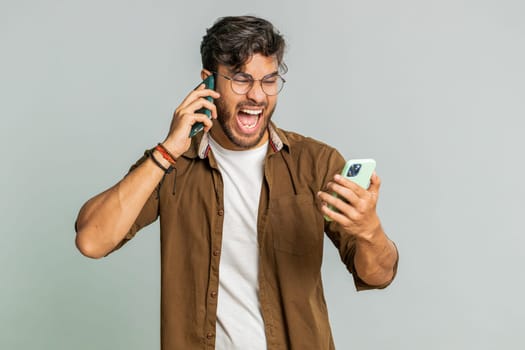Irritated nervous stressed Indian young man talking screaming on two mobile phones having conversation conflict quarrel complaint dispute discuss solve problem. Arabian guy isolated on gray background