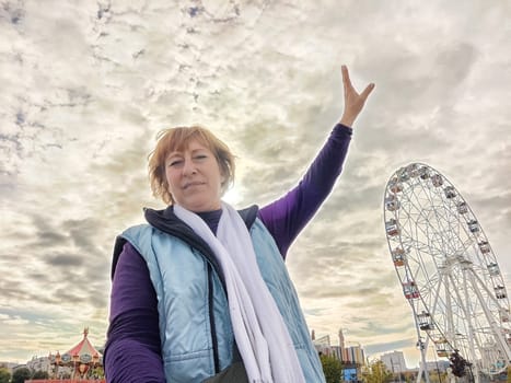 Relaxed mature caucasian female making selfie outside near big Ferris wheel. Positive senior woman against city street. Adult middle agged girl Blogger taking photo for internet