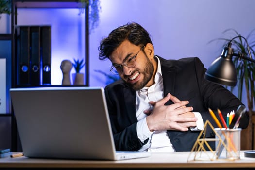 Stressed Indian businessman freelancer suffers from tension holding chest with heart pain attack risk of stroke symptom while working at modern home office workplace desk. Male Hindu in formal suit
