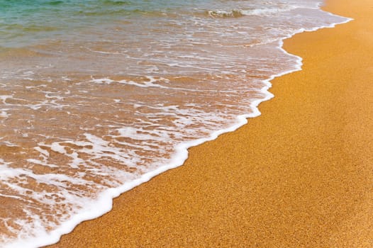 Background image of a soft blue ocean wave on a sandy beach. Close-up sea wave with white foam rolls onto golden sand.
