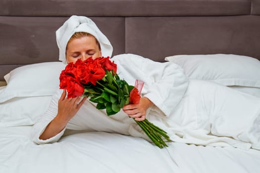 Tranquil romance: Beautiful young woman with long hair lying on a hotel bed holding a large bouquet of red roses, creating an atmosphere of elegance and romance.