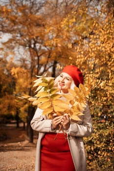 autumn woman in a red beret, a light coat and a red skirt, against the backdrop of an autumn park with yellow leaves in her hands.