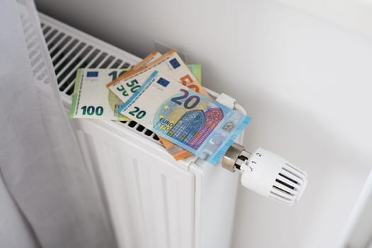 A heating radiator battery with a temperature controller from which 50 euro bills stick out. The concept of high heating costs. Rise in energy prices, energy crisis and recession. High quality photo