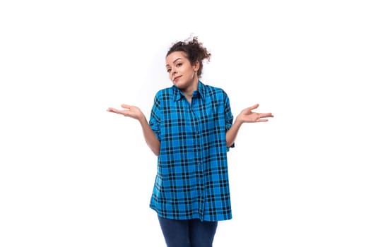 young curly brunette woman in a blue plaid shirt doubts and gesticulates against the background with copy space.