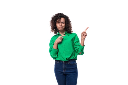 young caucasian brunette woman with curled hair dressed in a green shirt points her finger towards copy space. advertising concept.