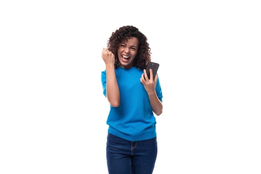 a slender young curly woman dressed in a blue summer jacket rejoices holding a mobile phone.