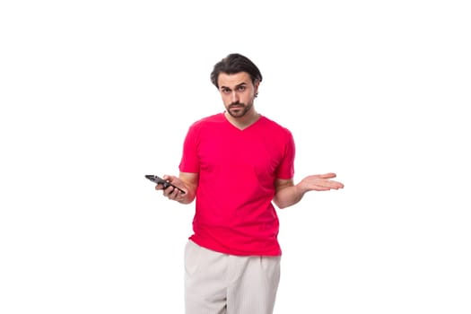 young well-groomed slender brunette man in a red T-shirt holds a phone in his hand and looks at the camera in confusion.