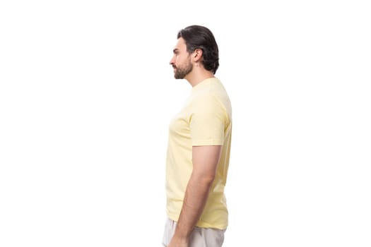 Caucasian brutal charming guy with a beard dressed in a summer T-shirt stands in profile.
