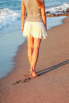 Young woman walking on wet sand beach lit by sunset light, view from back, detail on her calf leg muscles