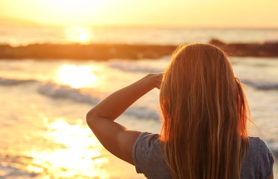 Sporty woman watches sunset over the sea, shading her eyes with one hand, view from behind only hair visible