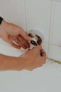 One young Caucasian guy, unrecognizable, wraps two hands of white tow on a faucet nut sticking out of the wall, standing bent over in the bathroom, close-up side view. Step by step.