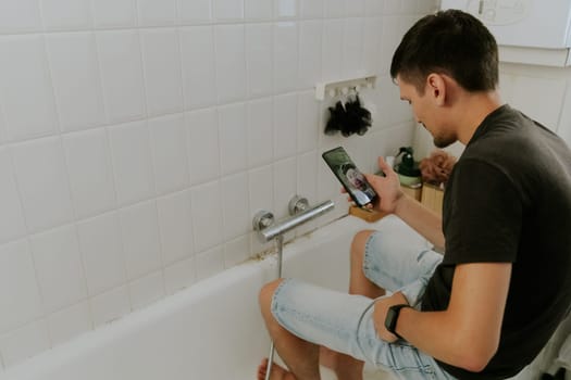 One young brunette Caucasian guy manually installs a faucet into the walls using an adjustable wrench while sitting on the edge of the bathtub, close-up side view. Step by step.