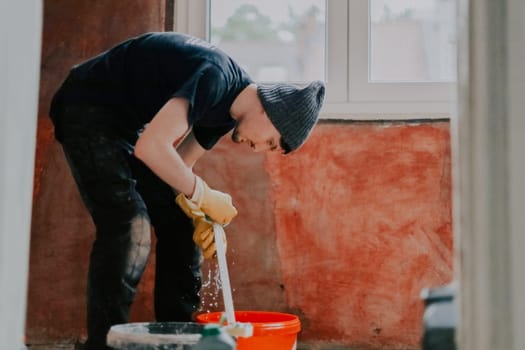 A young Caucasian man in black clothes with a hat on his head and yellow rubber gloves washes window frames in a bucket of water and a sponge, standing leaning in a room by the window, close-up side view.