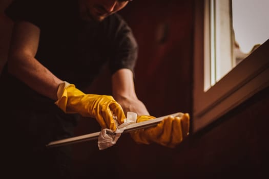 A young Caucasian man in black clothes and yellow rubber gloves is drying the plastic part of a window frame with a paper towel, standing in a room where renovations are underway, close-up view from below.