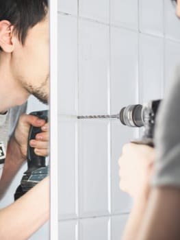 One young Caucasian man drills a hole between the seams of a tiled wall using a drill in the bathroom, reflected in the mirror of a wall cabinet, close-up side view.
