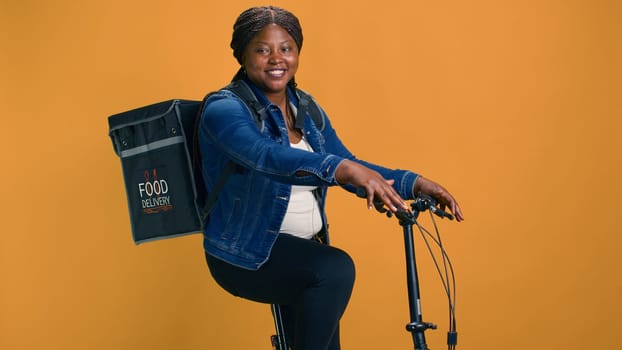 Reliable african american courier with bag on the back delivering food from restaurant to a local neighbourhood. Healthy black woman using bicycle as mode of transportation for efficient delivery.