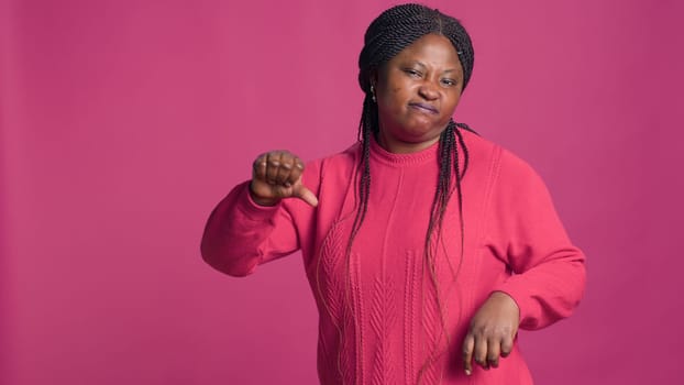 Expressive african american woman in pink sweater gesturing thumbs down towards camera. Fashion model displaying disapproval with her hands in front of bold isolated background.