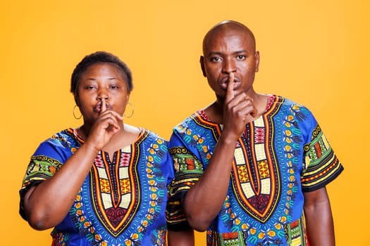 Confident black couple showing silence gesture while holding finger on lips and looking at camera. Serious man and woman asking to keep quiet portrait on studio orange background