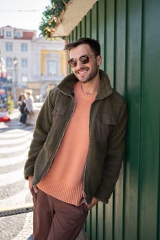 Man wear sunglasses standing outdoor autumn season. Portrait of stylish young adult guy wearing jacket looking at camera lean to wall in european city street on sunny day