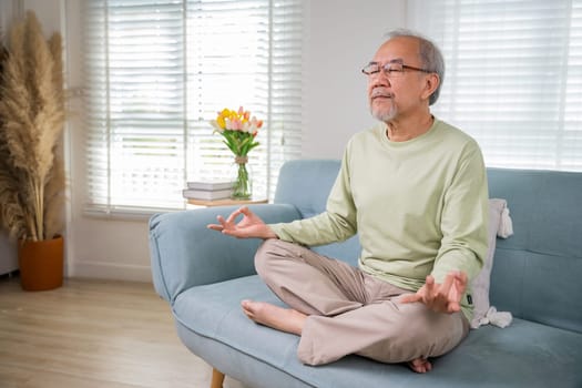 Asian old man practise yoga and meditation in lotus position and closed eyes, lifestyle senior man lotus pose doing yoga for mental balance breathing air relaxing on sofa at home, Healthy life habits