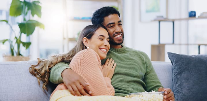 Love, hug and couple on watching tv on a sofa with popcorn for movie, film or streaming show at home. Relax, television and people embrace in a living room with cinema snack for comedy or series.