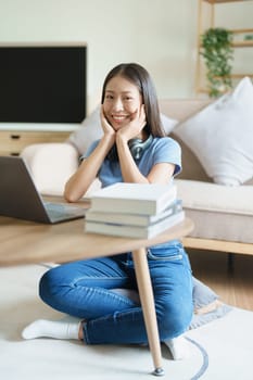 Portrait of young beautiful Asian woman showing smiling face during early morning online class with books, headphones and computer as study materials at home.