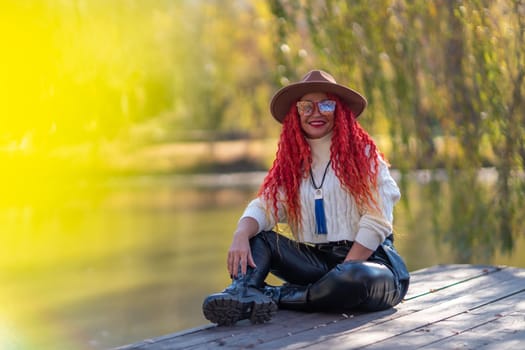 Autumn lake woman. In autumn, she sits by the pond on a wooden pier and admires nature with red hair and a hat. Tourism concept, weekend outside the city