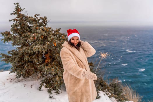 Outdoor winter portrait of happy smiling woman, light faux fur coat holding heart sparkler, posing against sea and snow background.