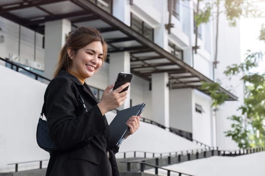 Attractive smiling Asian businesswoman wearing a suit standing in the city using an application on her mobile phone Read news on your smartphone fast connection Check out the outdoor mobile app.