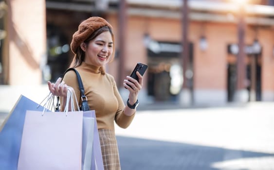 Beautiful Asian woman holding shopping bags, using smartphone and smiling while shopping on Black Friday outside the mall..