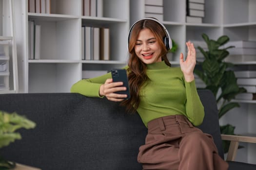 Happy Asian woman listening to music on mobile phone while sitting on sofa at home Relaxed smiling girl with headphones in the morning It's time to rest..