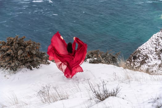 Woman red dress snow sea. Happy woman in a red dress in the snowy mountains by the emerald sea. The wind blows her clothes, posing against sea and snow background