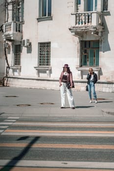 Woman city road crossing. Stylish woman in a hat crosses the road at a pedestrian crossing in the city. Dressed in white trousers and a jacket with a bag in her hands