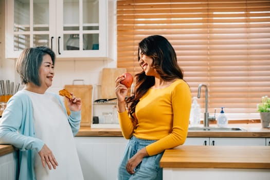 A cheerful Asian woman, with her mother, stands in the kitchen, holding an apple. Their warm embrace embodies the joy of teaching and learning within the family.