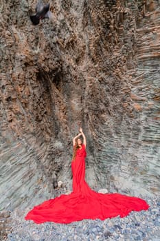woman sea red dress. Woman with long hair on a sunny seashore in a red flowing dress, back view, silk fabric waving in the wind. Against the backdrop of the blue sky and mountains on the seashore