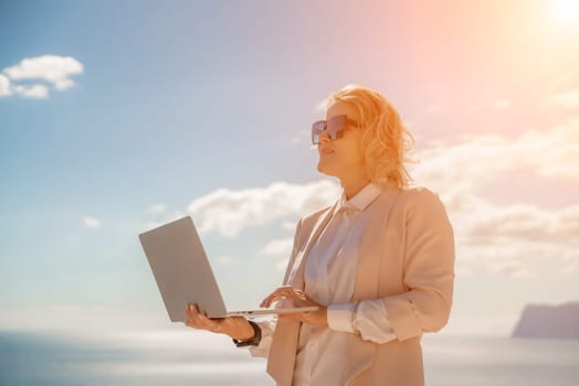 Freelance women sea. She is working on the computer. Good looking middle aged woman typing on a laptop keyboard outdoors with a beautiful sea view. The concept of remote work