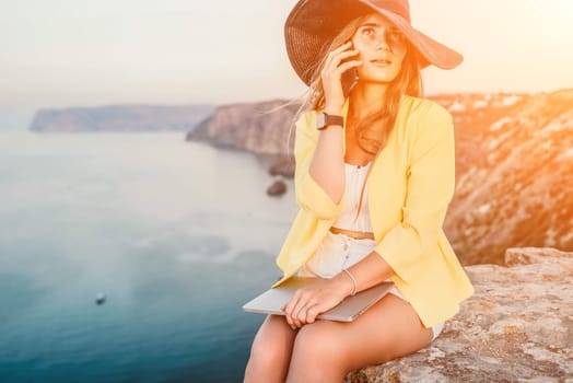 Successful business woman in yellow hat working on laptop by the sea. Pretty lady typing on computer at summer day outdoors. Freelance, travel and holidays concept.