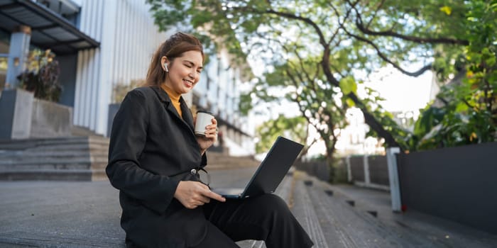 Smiling young Asian business woman using laptop sitting outdoor. hybrid working, searching job online thinking of digital solution.
