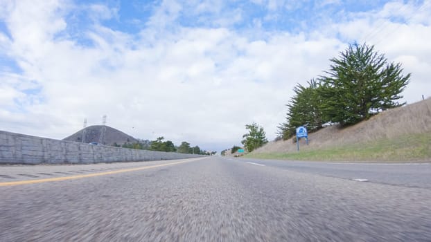 On a crisp winter day, a car cruises along the iconic Highway 101 near San Luis Obispo, California. The surrounding landscape is brownish and subdued, with rolling hills and patches of coastal vegetation flanking the winding road.