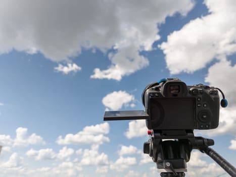black professional digital camera with flip screen on a tripod pointed at blue sky with white clouds at summer day