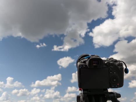 black professional digital camera on a tripod pointed at blue sky with white clouds at summer day