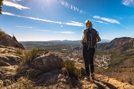 Beautiful mountain standing young woman with backpack on top at sunset in summer. Landscape with a sporty girl, hills, blue sky with sun rays. Travel and tourism. High quality photo